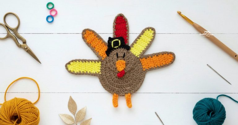 Crochet turkey applique centered on a white wooden background with scissors, a crochet hook, yarn, a yarn needle, stitch markers, and fall-inspired decor nearby.
