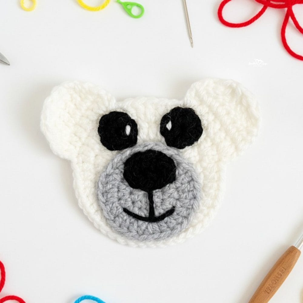 White crochet bear applique with a gray snout, black eyes, and black nose on a white background with yarn, a crochet hook, and colorful stitch markers nearby.