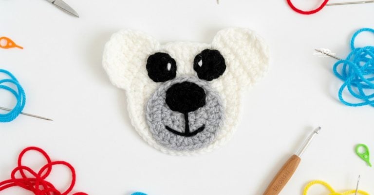 Crochet bear face applique in white and gray centered on a white desktop with yarn, a crochet hook, scissors, and yarn needles around the edges.