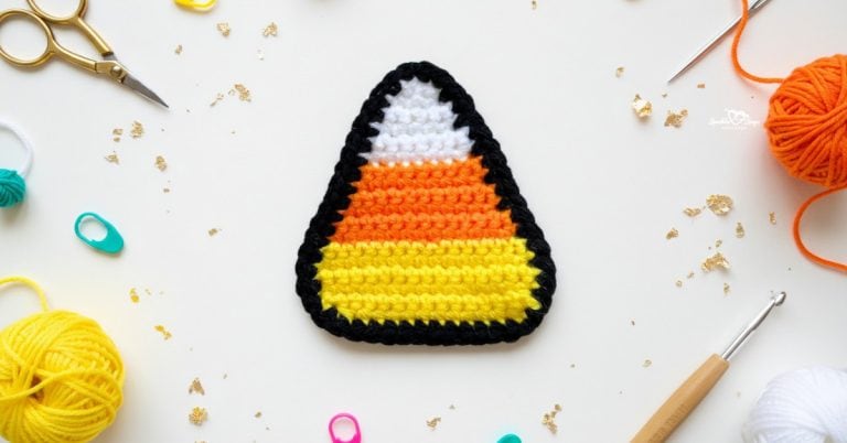 Crochet candy corn applique in white, orange, and yellow with a black border centered on a white desktop with yarn, a crochet hook, scissors, stitch markers, and gold confetti around it.
