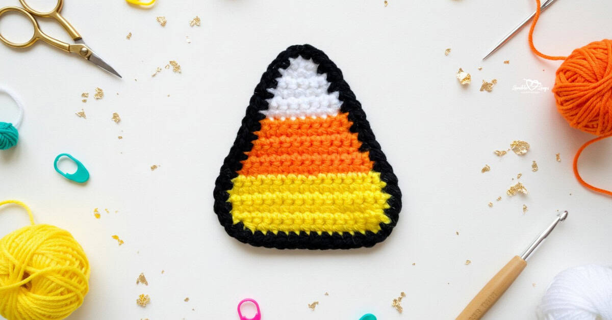 Crochet candy corn applique in white, orange, and yellow with a black border centered on a white desktop with yarn, a crochet hook, scissors, stitch markers, and gold confetti around it.