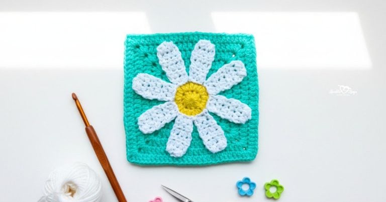Overhead view of a turquoise crochet granny square with a white daisy and yellow center on a white desk, styled with a crochet hook, yarn, scissors, and flower-shaped stitch markers.
