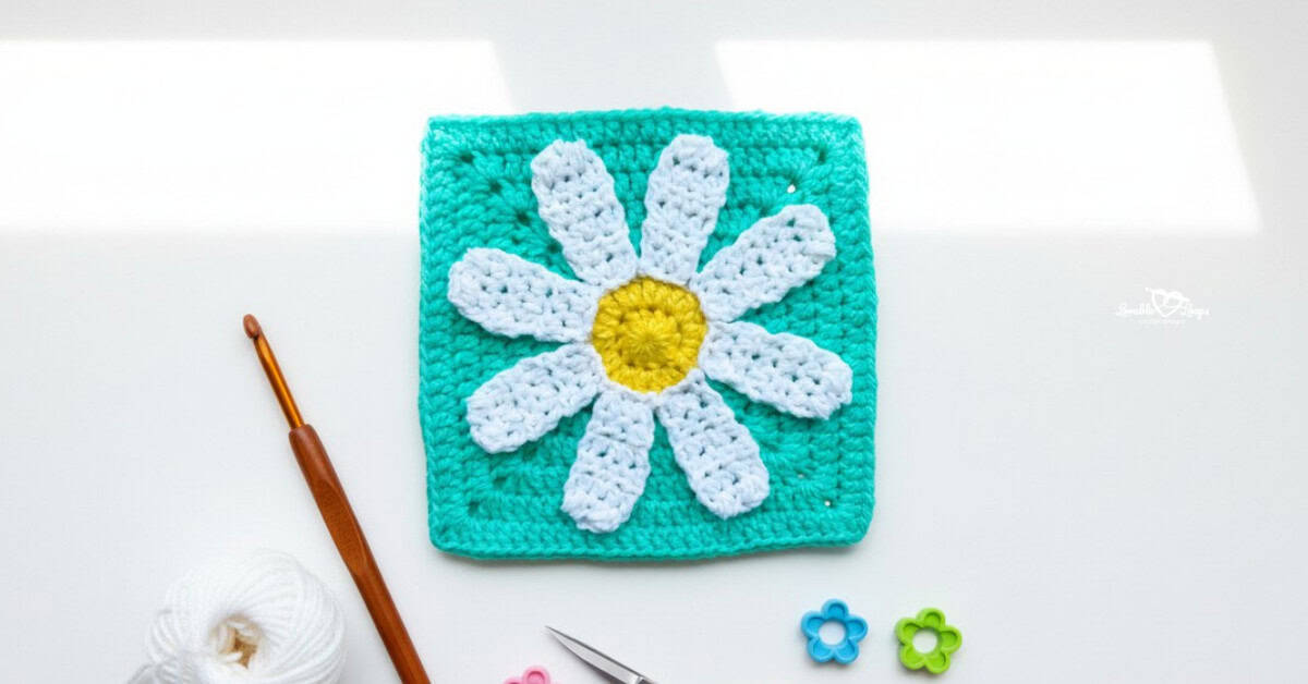 Overhead view of a turquoise crochet granny square with a white daisy and yellow center on a white desk, styled with a crochet hook, yarn, scissors, and flower-shaped stitch markers.
