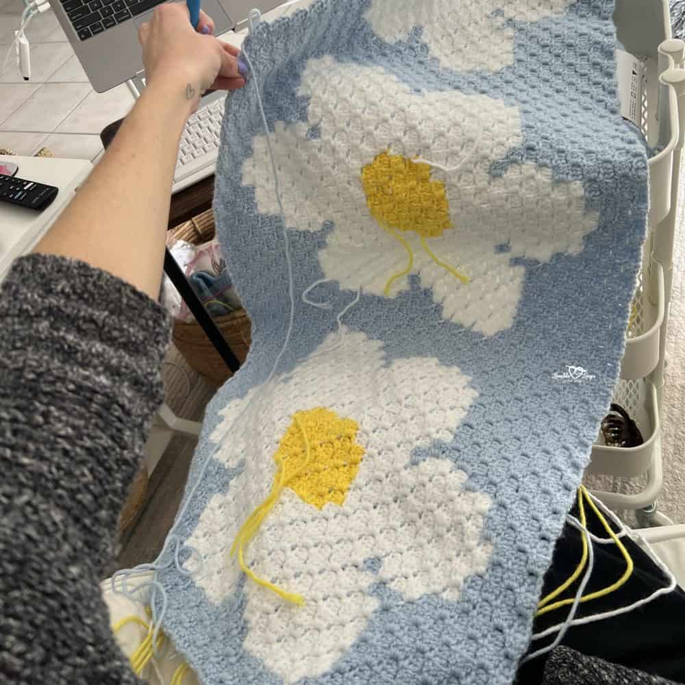 Crochet daisy table runner in progress with white daisies and yellow centers on a blue background, shown being worked on by hand.