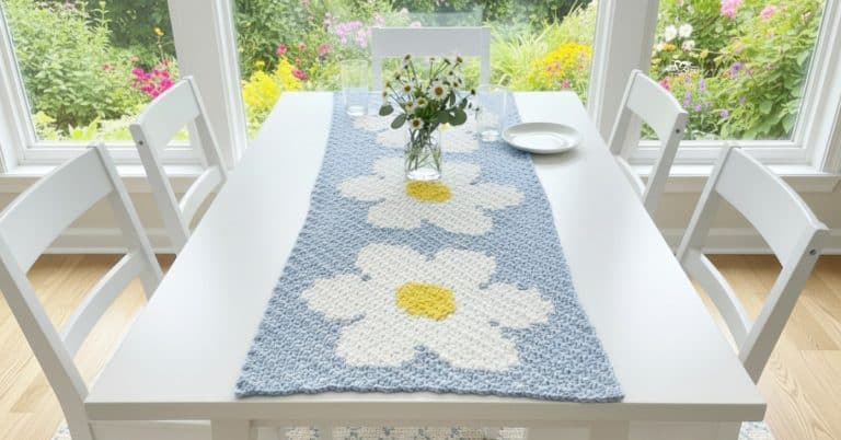 Crochet daisy table runner with large white daisies and yellow centers displayed on a white dining table with chairs and a vase of fresh flowers.