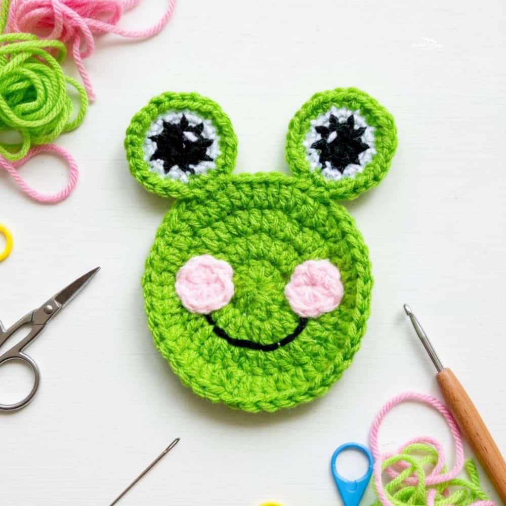 Cute crochet frog applique in bright green yarn with big eyes, pink cheeks, and a stitched smile on a white background with crochet tools nearby.