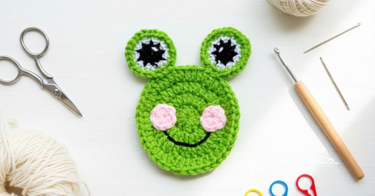 Green crochet frog face applique with large eyes and pink cheeks centered on a white desktop with scissors, a crochet hook, yarn, and stitch markers around the edges.