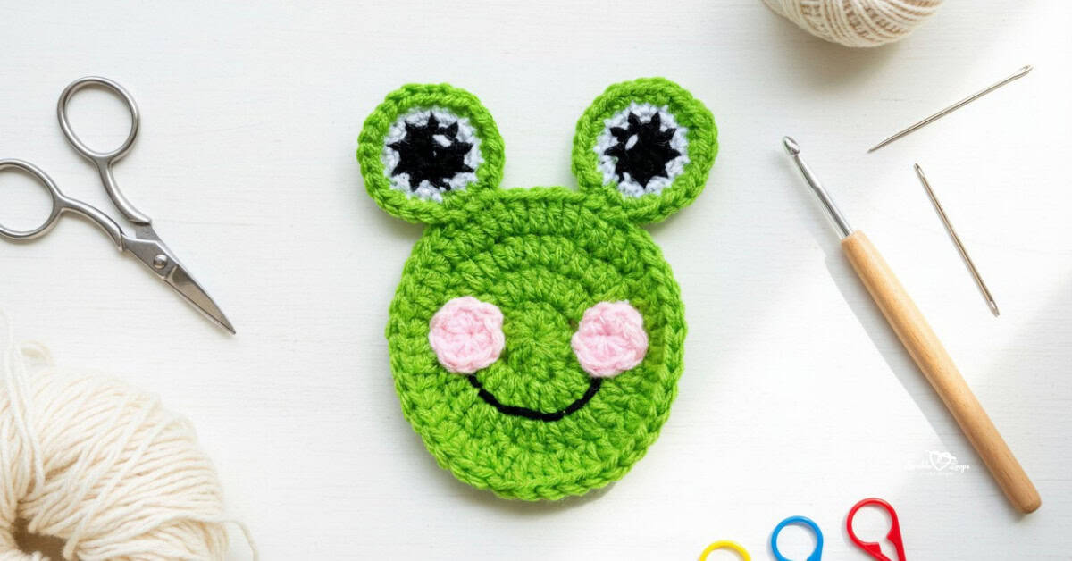 Green crochet frog face applique with large eyes and pink cheeks centered on a white desktop with scissors, a crochet hook, yarn, and stitch markers around the edges.