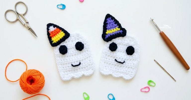 Two crochet ghost appliques on a white desktop, one wearing a candy corn hat and one wearing a purple witch hat, with scissors, a crochet hook, orange yarn, and colorful stitch markers nearby.