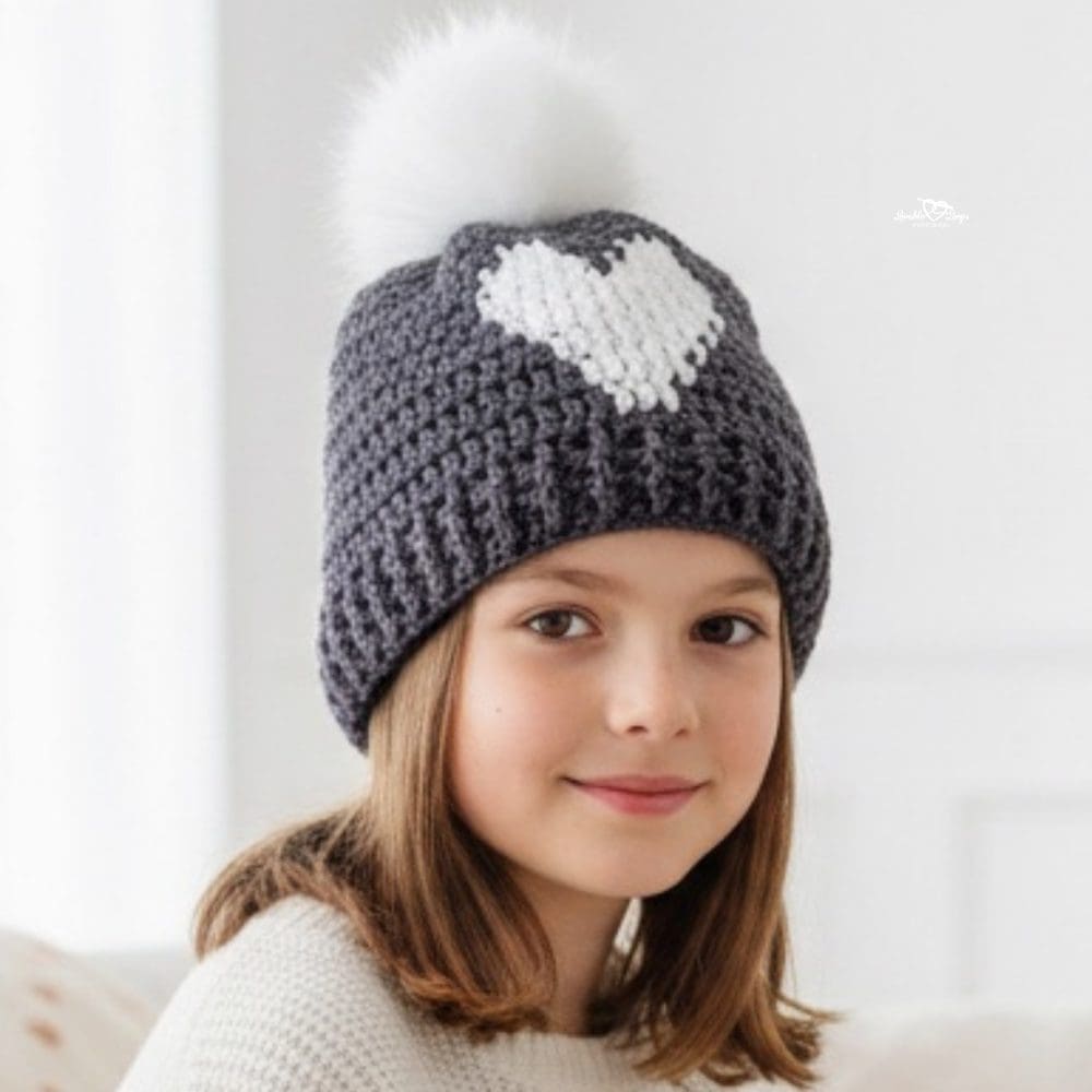 Child wearing a charcoal gray crochet beanie with a white heart design and a white pom pom.