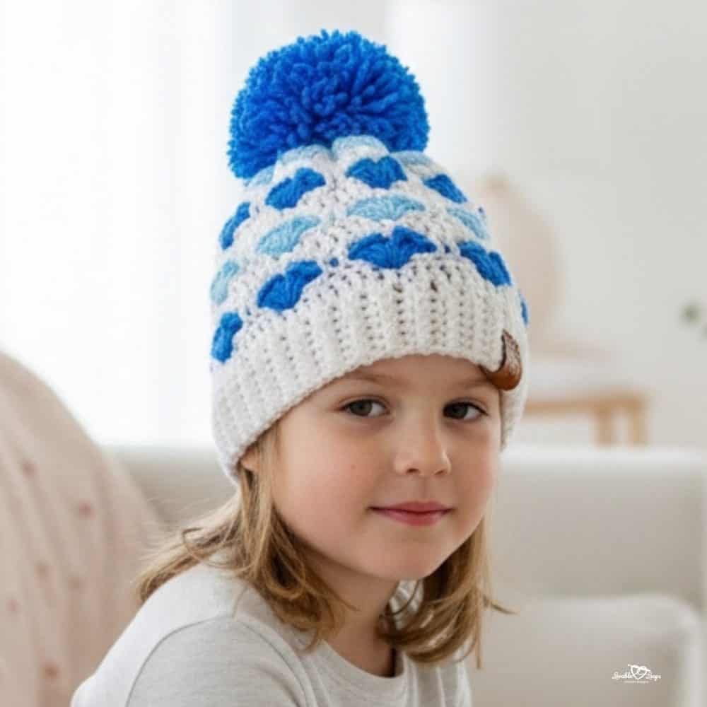 Child wearing a white crochet beanie with blue heart= stitches and a blue pom pom.