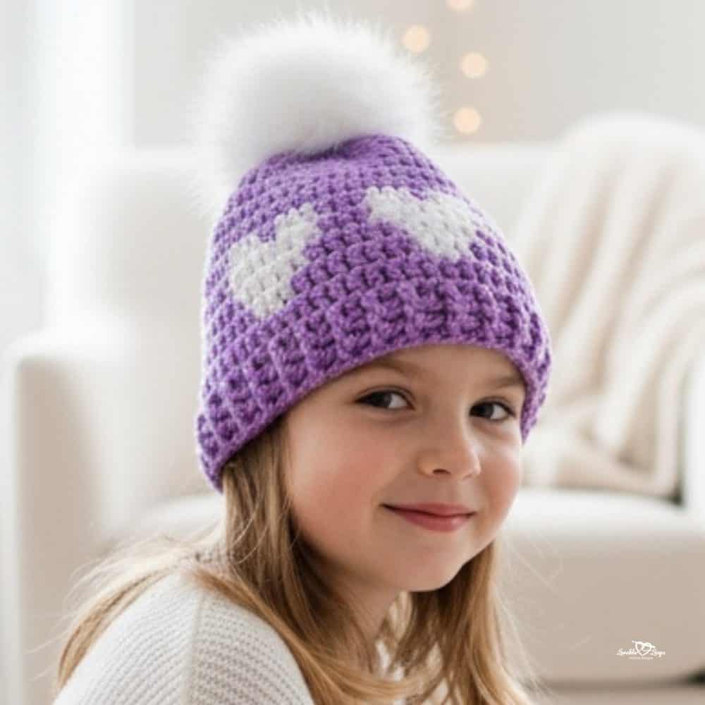 Child wearing a purple crochet beanie with two white heart designs and a white pom pom.
