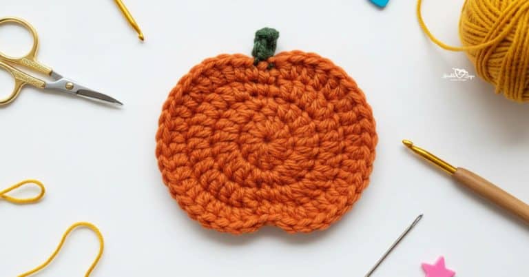 Orange crochet pumpkin applique with a small green stem centered on a white desktop with scissors, a crochet hook, yarn, a yarn needle, and small colorful craft details around it.