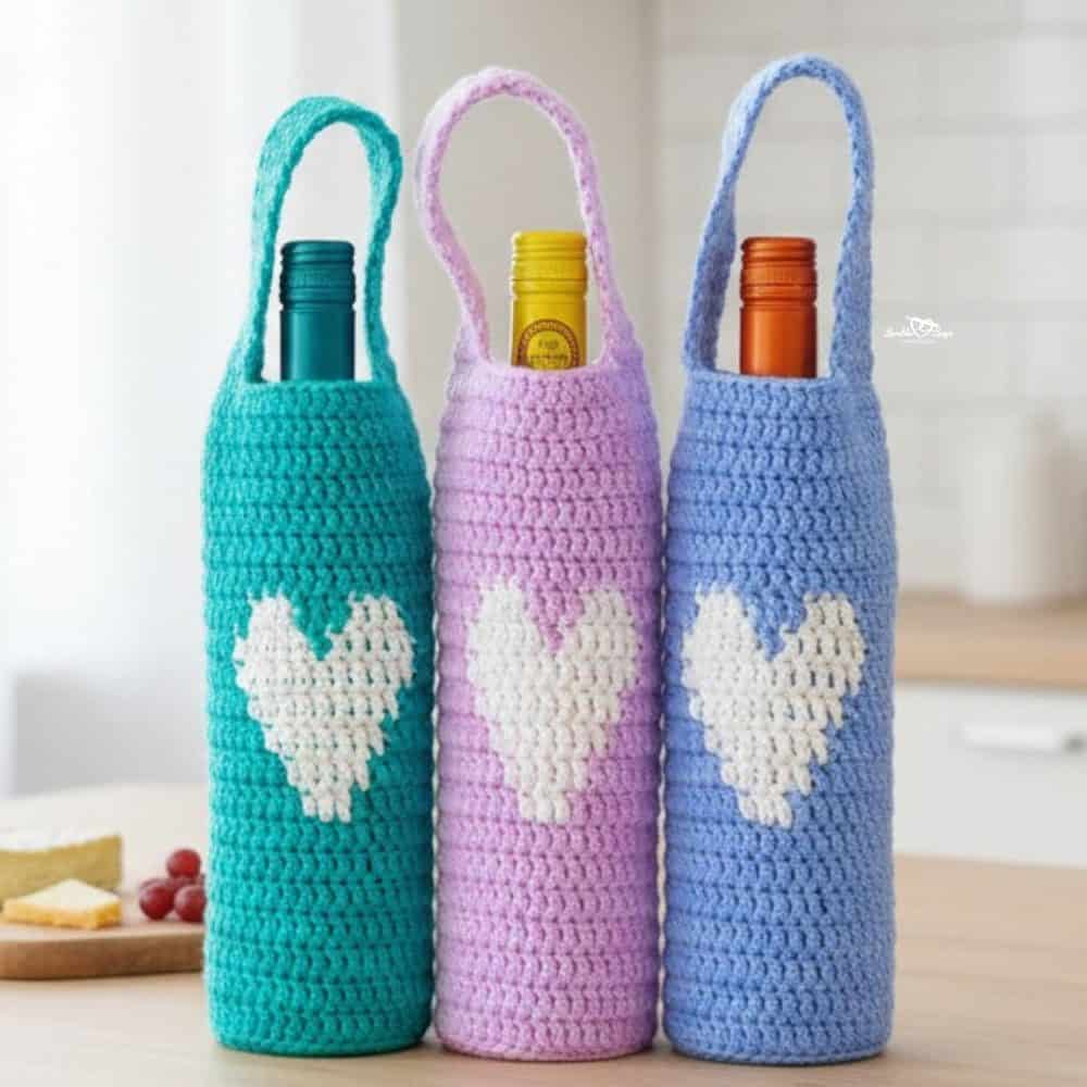 Three crochet wine bottle holders with white heart motifs in teal, lavender, and periwinkle displayed together on a light wood kitchen island with simple giftable decor.