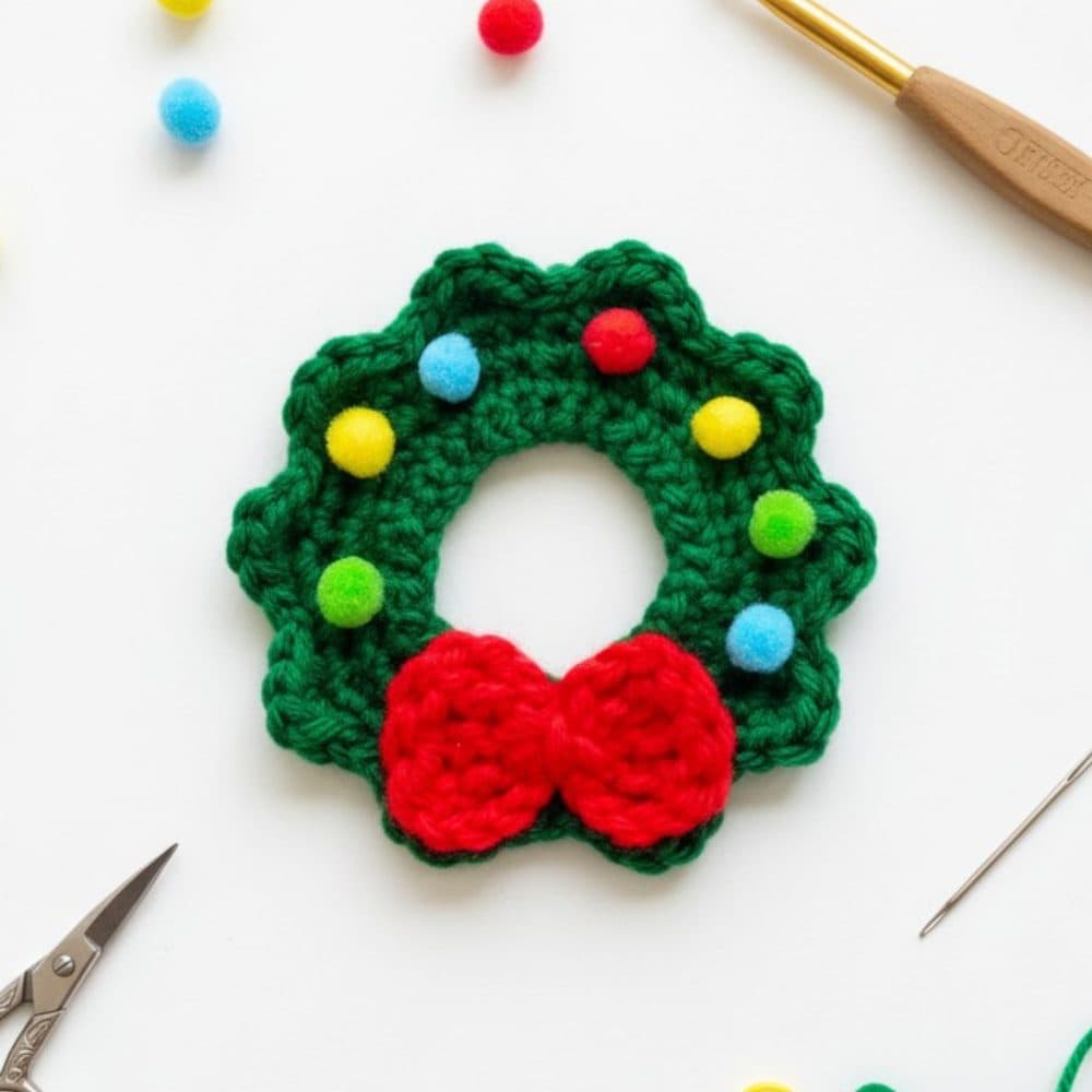 Small crochet Christmas wreath applique in green yarn with a bright red bow and colorful pom-pom accents on a white background.