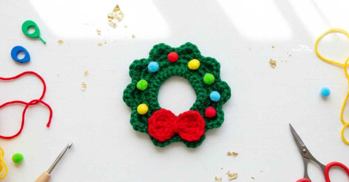 Crochet wreath applique centered on a white desktop with red and yellow yarn, scissors, a crochet hook, colorful stitch markers, and festive gold accents around it.