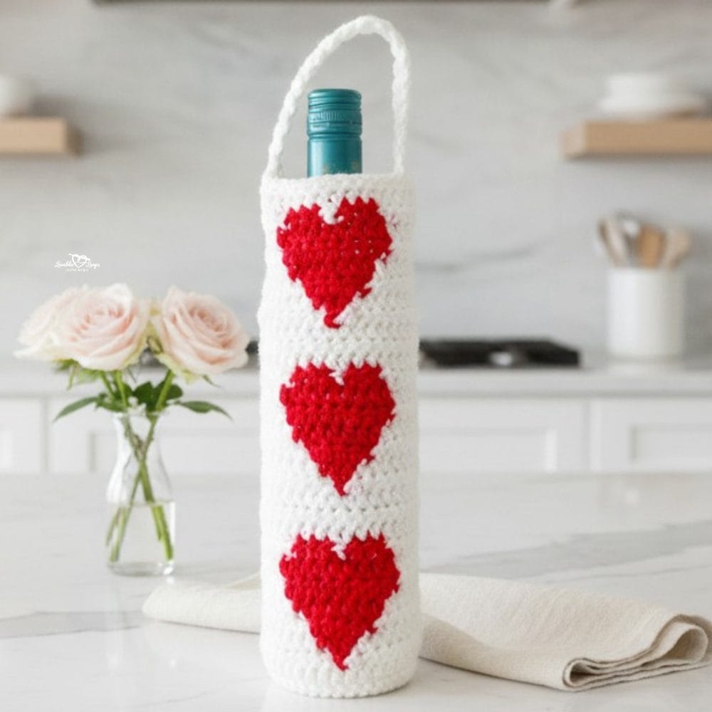 Crochet wine bottle bag with red heart motifs displayed on a bright kitchen island with soft floral accents and a clean modern white kitchen background.