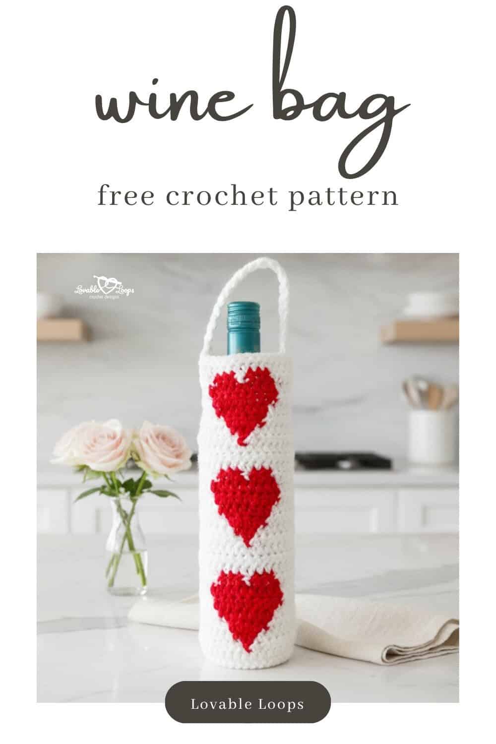 White crochet wine bottle bag with three red heart motifs displayed on a white kitchen island beside pale pink roses and a cream linen napkin.
