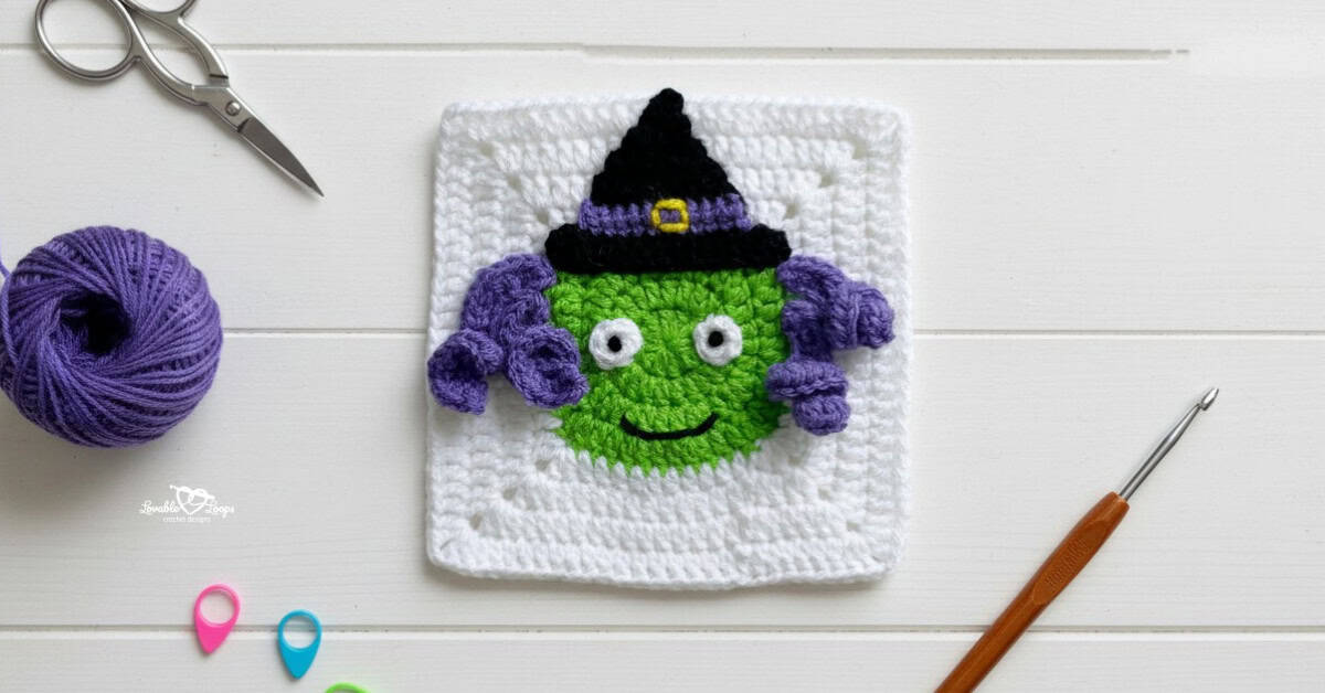 Overhead view of a white crochet granny square with a green witch face applique, curly purple hair, and a black witch hat, styled on a white wood surface with a crochet hook, scissors, purple yarn, and stitch markers.