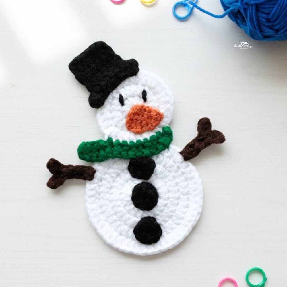Crochet snowman applique in white yarn with a black hat, green scarf, orange nose, black buttons, and brown stick arms on a white desktop with yarn and colorful stitch markers nearby.