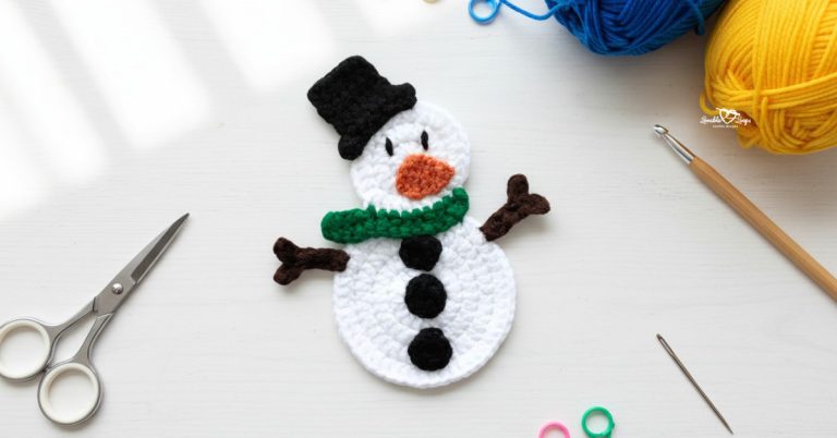 Crochet snowman applique centered on a white desktop with scissors, a crochet hook, yarn, a yarn needle, and colorful stitch markers around the edges.