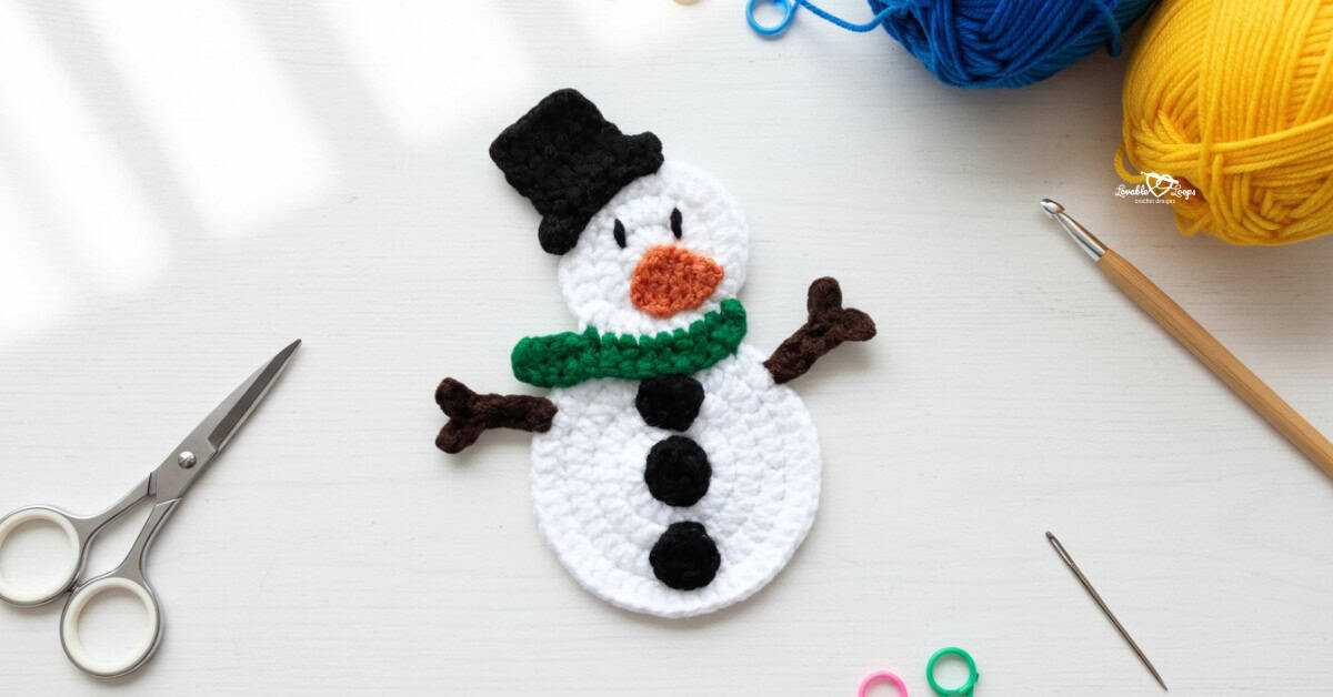 Crochet snowman applique centered on a white desktop with scissors, a crochet hook, yarn, a yarn needle, and colorful stitch markers around the edges.
