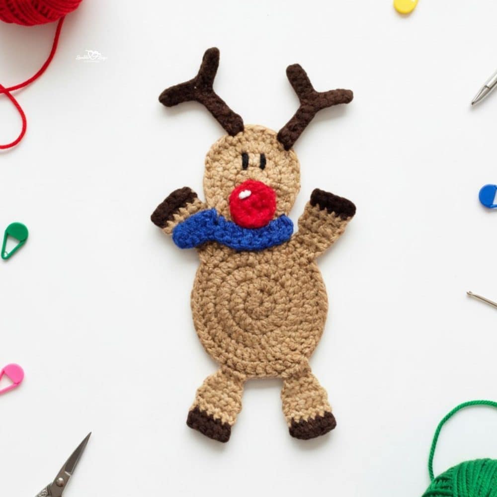 Crochet reindeer applique in tan and brown yarn with dark antlers, a bright red nose, and a blue scarf on a white desktop with scissors, yarn, a yarn needle, and colorful stitch markers nearby.