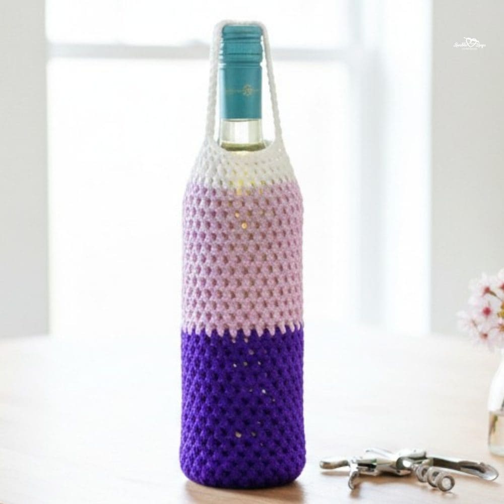 Crochet wine bottle holder with a white top, light pink center, and deep purple base standing on a light wood kitchen island beside a small vase of pink flowers and a corkscrew.