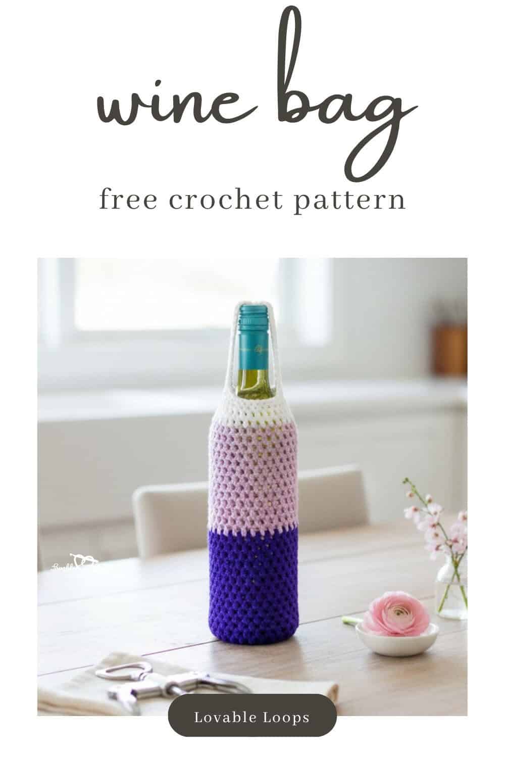 Crochet wine bottle bag with a white top, soft pink center, and deep purple base displayed on a light wood table with a small white dish, pink flower, tiny vase, and corkscrew nearby.