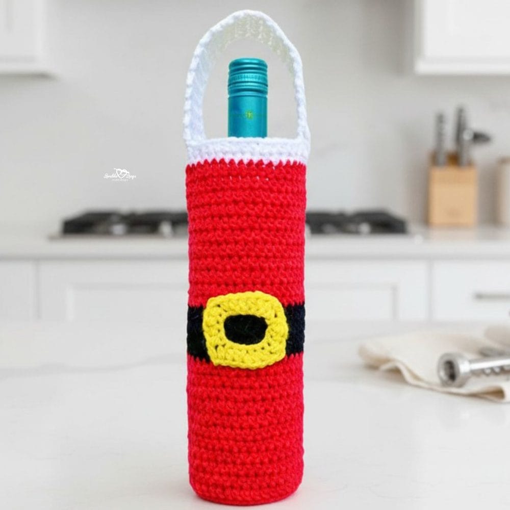 Santa crochet wine bottle holder standing on a white kitchen island, styled with wooden trees, a cream napkin, and a corkscrew in a bright holiday kitchen.