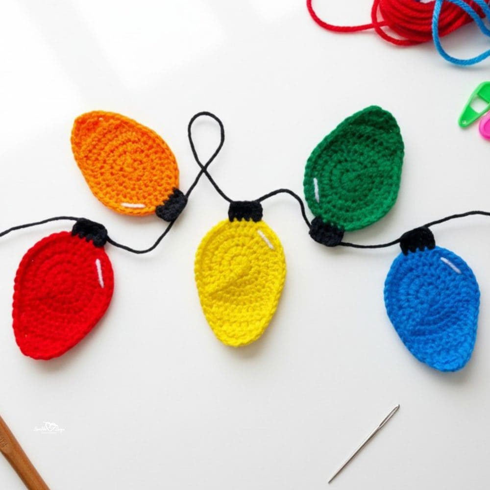 Colorful crochet Christmas light appliques in red, orange, yellow, green, and blue connected by black yarn on a white background.