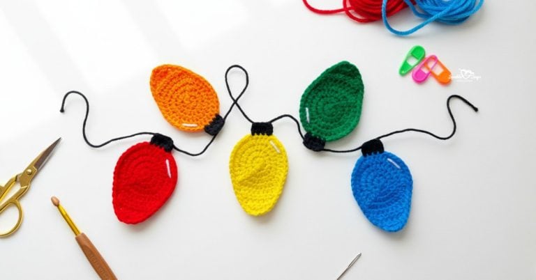 Crochet Christmas lights applique garland in red, orange, yellow, green, and blue on a white desktop with scissors, a crochet hook, yarn, stitch markers, and a yarn needle nearby.