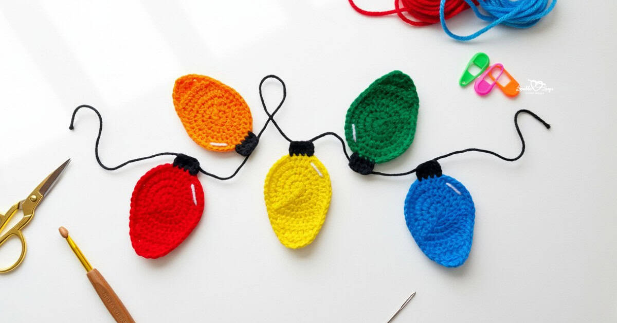 Crochet Christmas lights applique garland in red, orange, yellow, green, and blue on a white desktop with scissors, a crochet hook, yarn, stitch markers, and a yarn needle nearby.