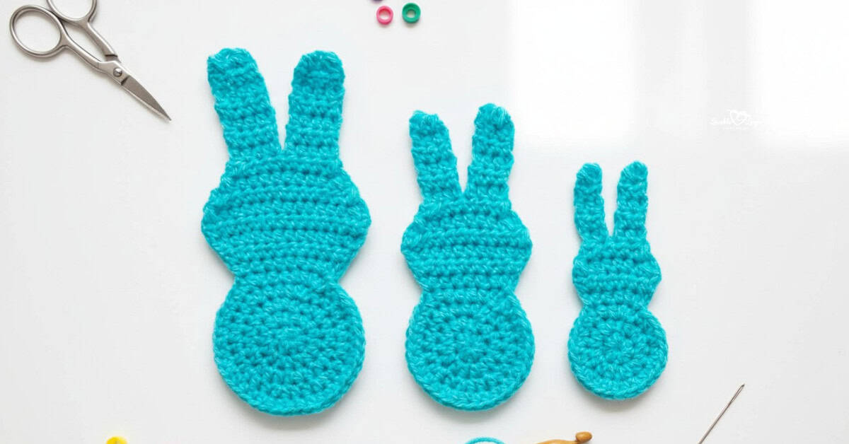 Three turquoise crochet Peeps bunny appliques in different sizes arranged on a white desktop with scissors, a crochet hook, and stitch markers nearby.