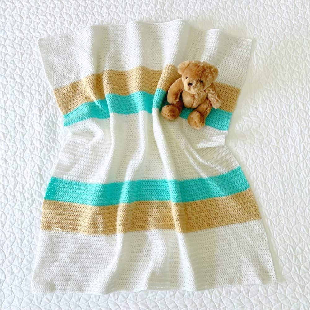 Neutral crochet baby blanket with white, cream, and aqua stripes laid out on a white quilted bedspread with a teddy bear.