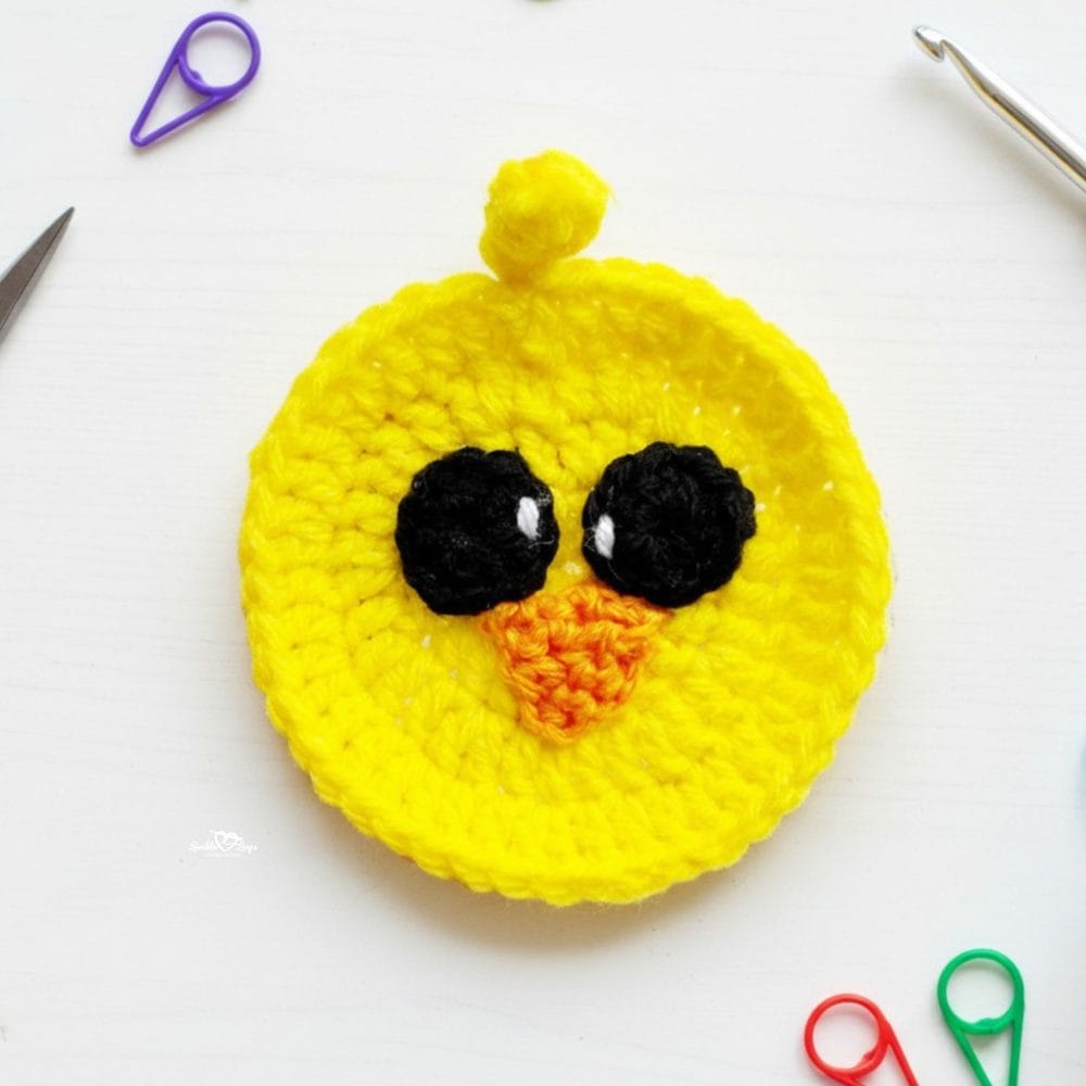 Crochet chick applique in bright yellow yarn with black eyes and an orange beak on a white desktop with colorful stitch markers and a crochet hook nearby.