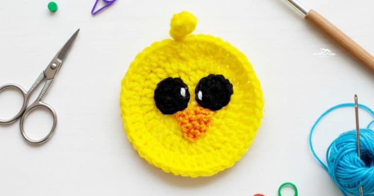 Yellow crochet chick applique centered on a white desktop with scissors, a crochet hook, blue yarn, a yarn needle, and colorful stitch markers around the edges.