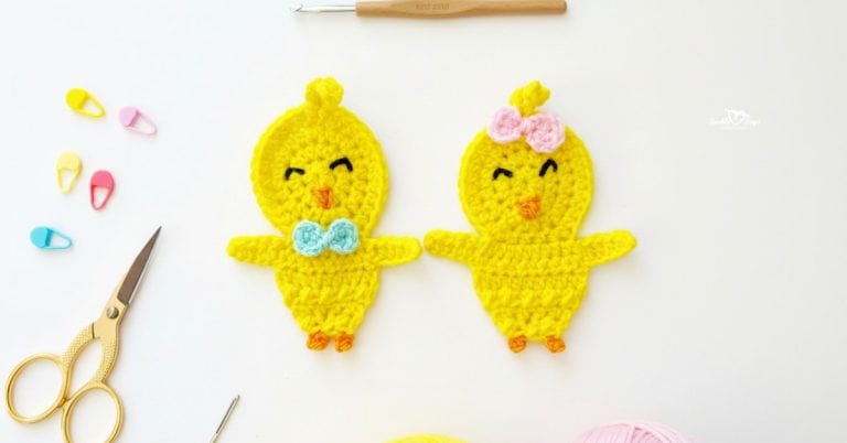 Two crochet chick appliques in bright yellow yarn centered on a white desktop with scissors, a crochet hook, yarn, and colorful stitch markers nearby.