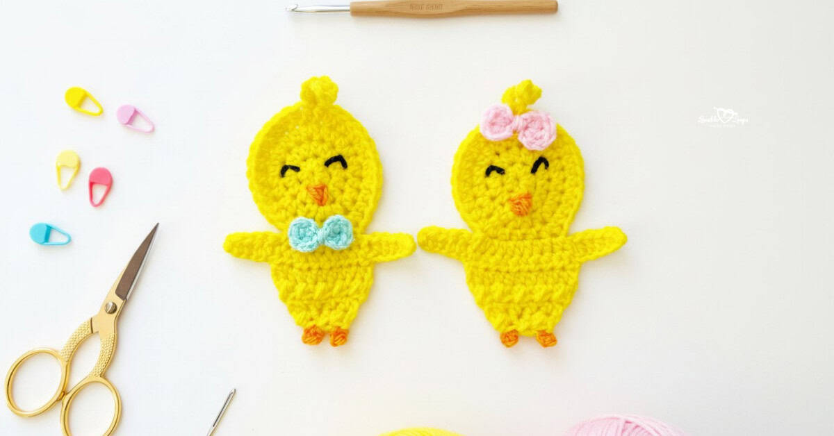 Two crochet chick appliques in bright yellow yarn centered on a white desktop with scissors, a crochet hook, yarn, and colorful stitch markers nearby.