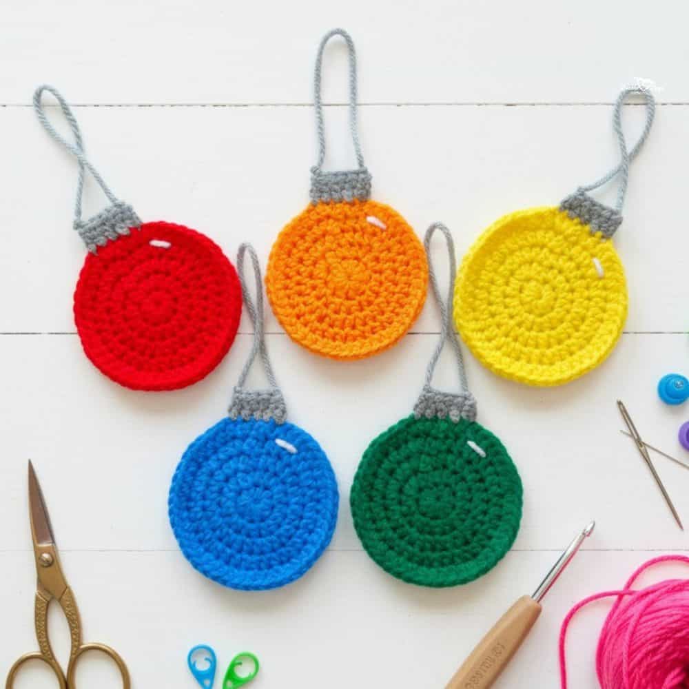Five crochet ornament appliques in red, orange, yellow, blue, and green with gray tops and hanging loops on a white background.