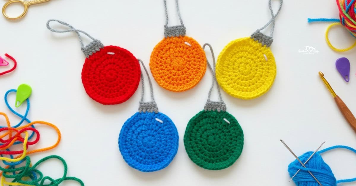 Colorful crochet ornament appliques arranged on a white desktop with yarn, scissors, a crochet hook, yarn needles, and stitch markers nearby.