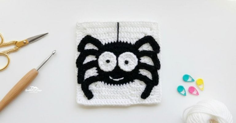 Overhead view of a white crochet granny square with a black spider applique, styled on a white desk with gold scissors, a crochet hook, white yarn, and colorful stitch markers.