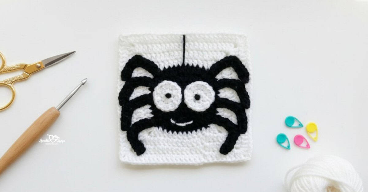 Overhead view of a white crochet granny square with a black spider applique, styled on a white desk with gold scissors, a crochet hook, white yarn, and colorful stitch markers.