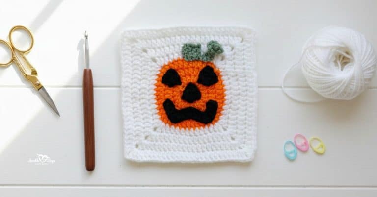 Overhead view of a white crochet granny square with an orange jack-o-lantern face applique, styled on a white wood surface with gold scissors, a crochet hook, white yarn, and pastel stitch markers.