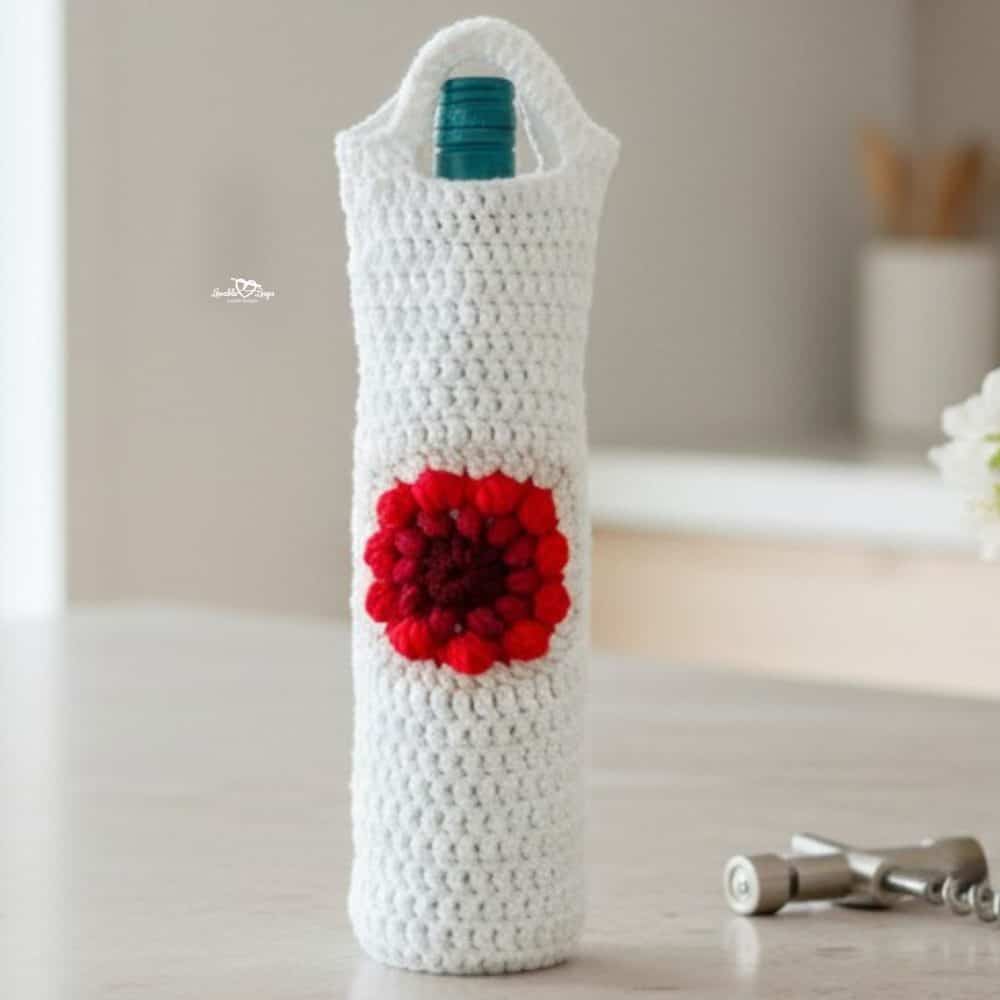 White crochet wine bottle holder with a red flower motif standing on a light kitchen island, styled with a small glass vase of white flowers, a corkscrew, and cream linen.