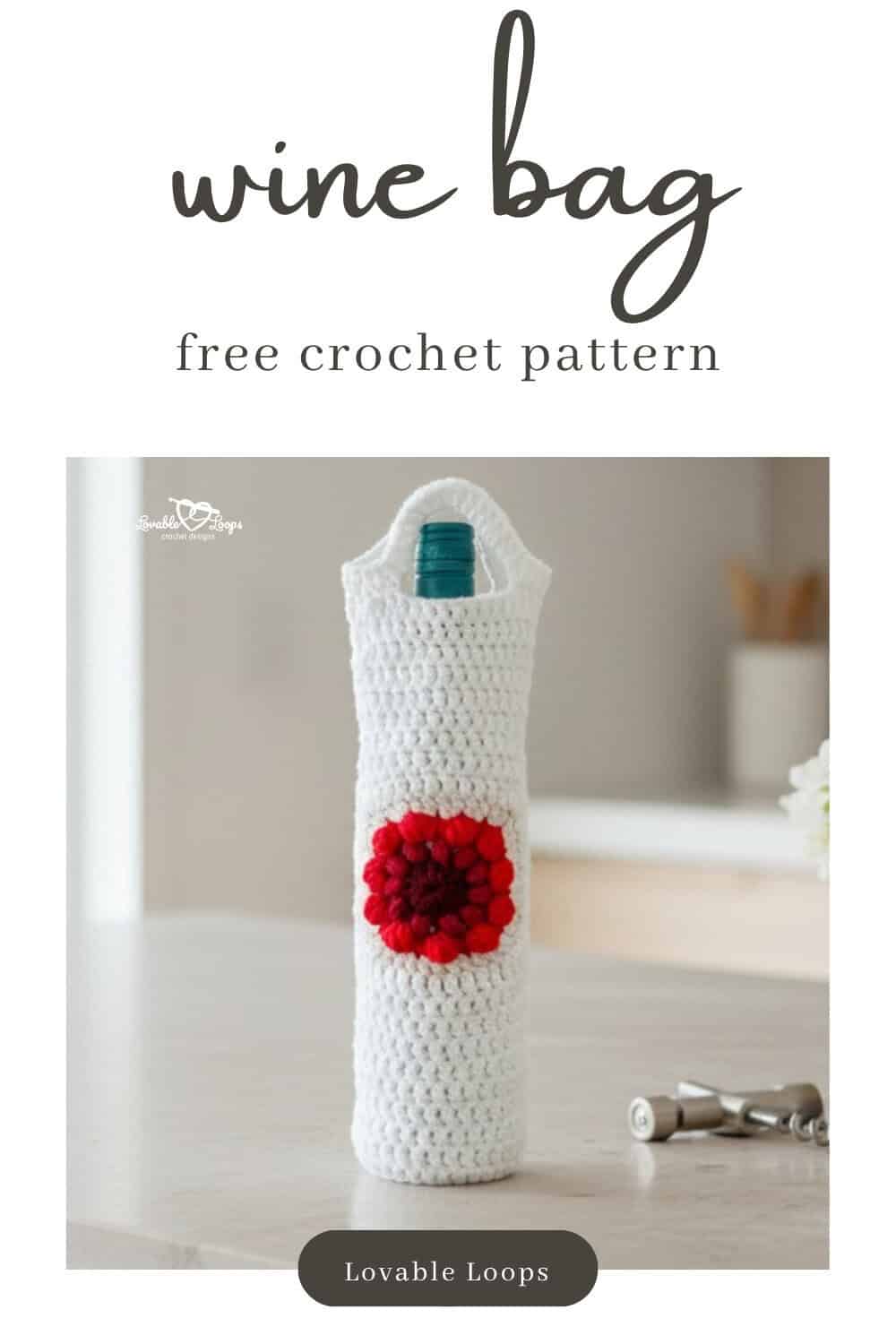 White crochet wine bottle bag with a red flower motif displayed on a light kitchen island beside a corkscrew, cream linen, and a small vase of white flowers.