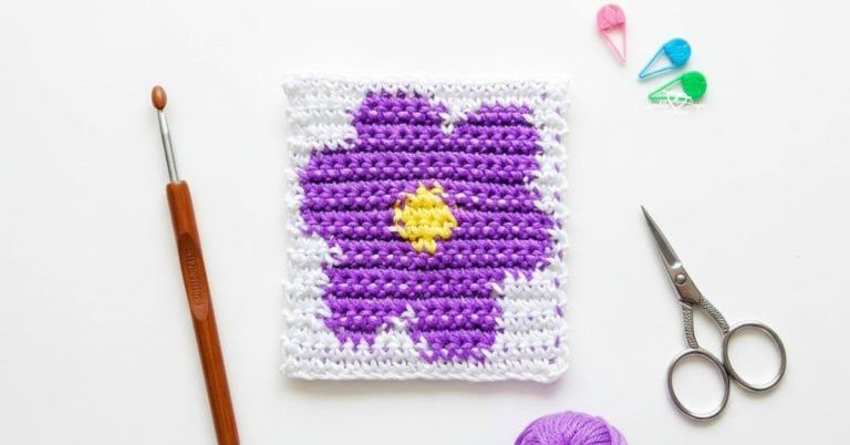 Overhead view of a white crochet square with a purple flower and yellow center on a white desk, styled with a crochet hook, small scissors, purple yarn, and colorful stitch markers.