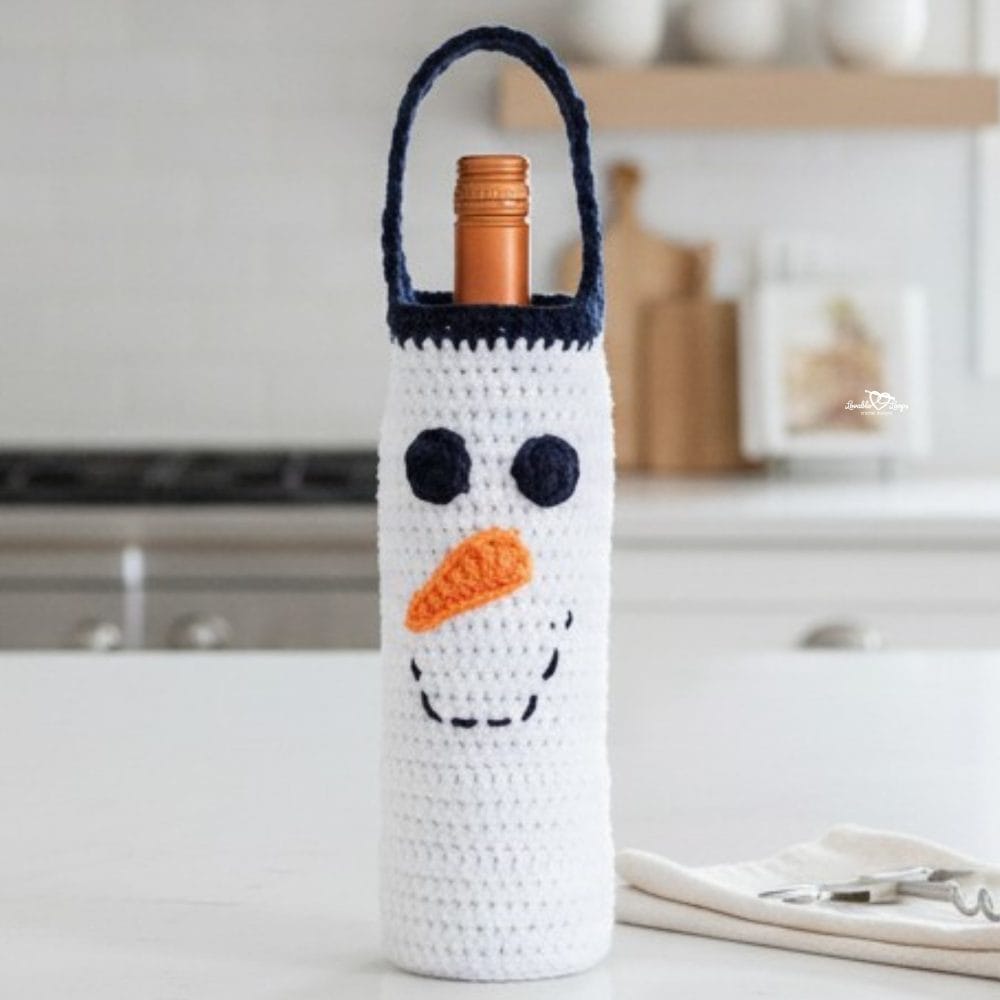 Snowman crochet wine bottle holder standing on a white kitchen island, styled with wooden trees, a cream napkin, and a corkscrew in a bright modern holiday kitchen.