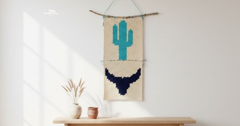 Southwestern crochet wall hanging with a turquoise cactus and navy bull skull motif displayed on a bright neutral wall above a light wood bench with simple pottery and dried stems.