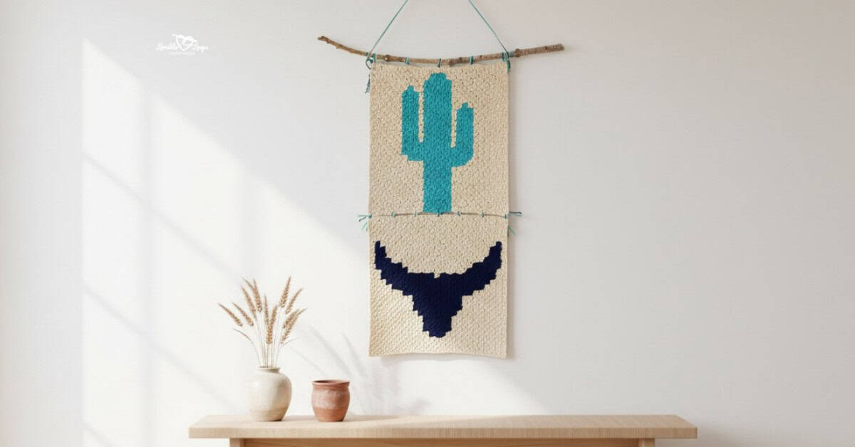Southwestern crochet wall hanging with a turquoise cactus and navy bull skull motif displayed on a bright neutral wall above a light wood bench with simple pottery and dried stems.